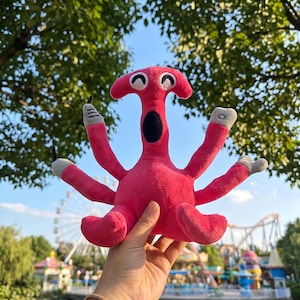 Puede incluir: Un peluche de color rosa brillante con una cabeza en forma de martillo, una boca ovalada negra y cuatro brazos largos con puntas grises. La criatura tiene dos ojos blancos con pupilas negras curvas y dos piernas cortas en la base del cuerpo.