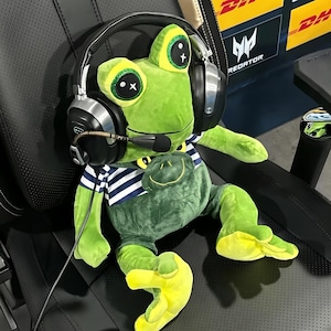 May include: A bright green plush frog toy wearing a blue and white striped shirt and large black over-ear gaming headphones with a microphone. The frog has large, glittery eyes with white star accents and yellow feet, sitting upright on a black chair.