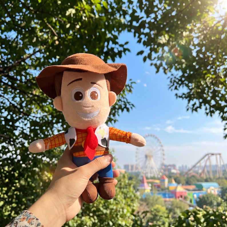 May include: A plush toy character wearing a brown cowboy hat, a yellow and orange plaid shirt, a red neckerchief, a white vest with black cow-print spots, and blue pants. The character has embroidered facial features and brown boots.