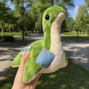 May include: A plush toy shaped like a long-necked creature with green fur, a cream-coloured underside, and black button eyes. The toy features a blue fabric patch on its side and a green and white checkered patch, both attached with visible red stitching.