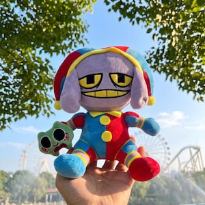 May include: A plush toy featuring a purple character with a wide grin and yellow eyes, wearing a red and blue jester hat and matching outfit with yellow buttons. The character holds a small, light green, round plush with two large black eyes.