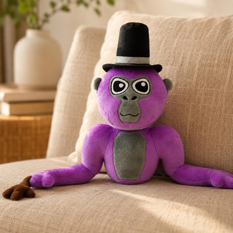 May include: A plush purple gorilla toy featuring a black top hat with a grey band. The gorilla has a grey face, chest patch, and ears, with large white eyes and black pupils. One arm extends to the left with a brown hand, while the other is tucked.