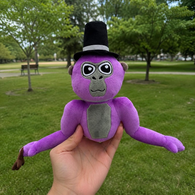 May include: A plush purple gorilla toy featuring a grey chest patch and face, wearing a black top hat with a white band. The stuffed animal has large white eyes with black pupils and long arms extending outward, crafted from soft fabric.
