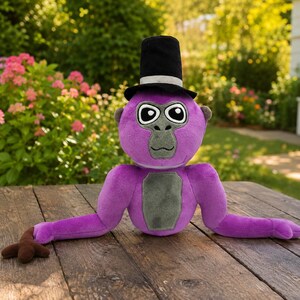 May include: A plush purple gorilla toy featuring a black top hat with a silver band, a gray face, and a gray chest patch. The stuffed animal has large white eyes with black pupils and long arms extending outward on a wooden surface.