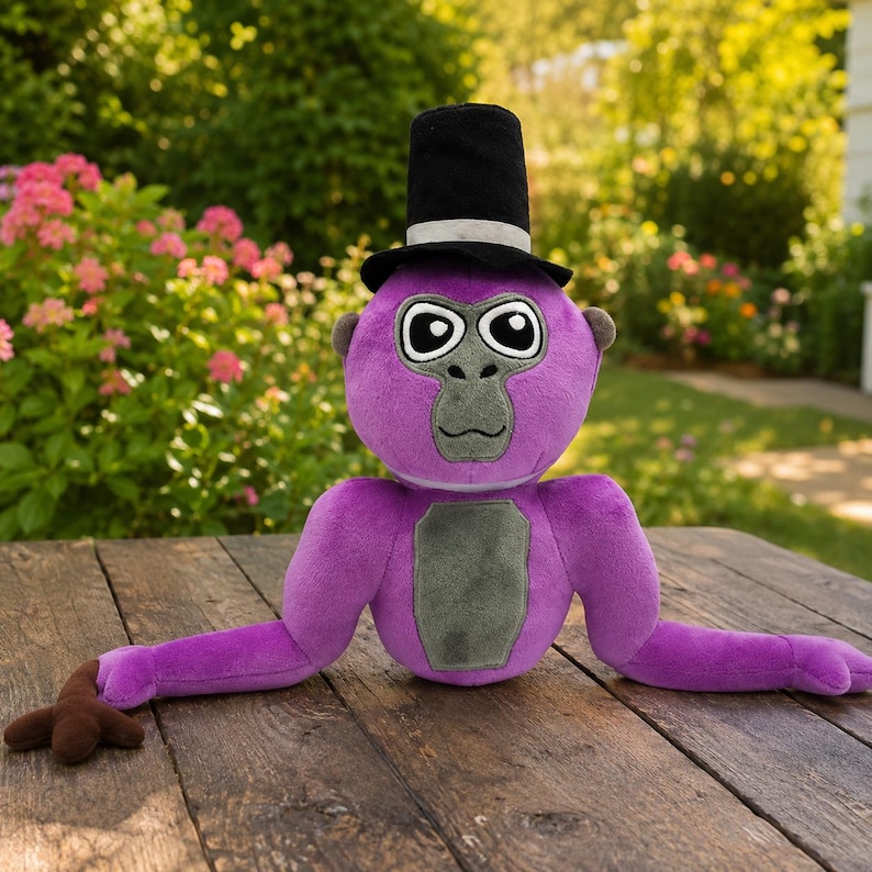 May include: A plush purple gorilla toy featuring a black top hat with a silver band, a gray face, and a gray chest patch. The stuffed animal has large white eyes with black pupils and long arms extending outward on a wooden surface.