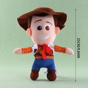 May include: A plush toy character wearing a brown cowboy hat, a yellow and orange plaid shirt, a white cow-print vest, a red neckerchief, and blue pants. The toy measures 9.84 inches in height as indicated by the vertical measurement line.