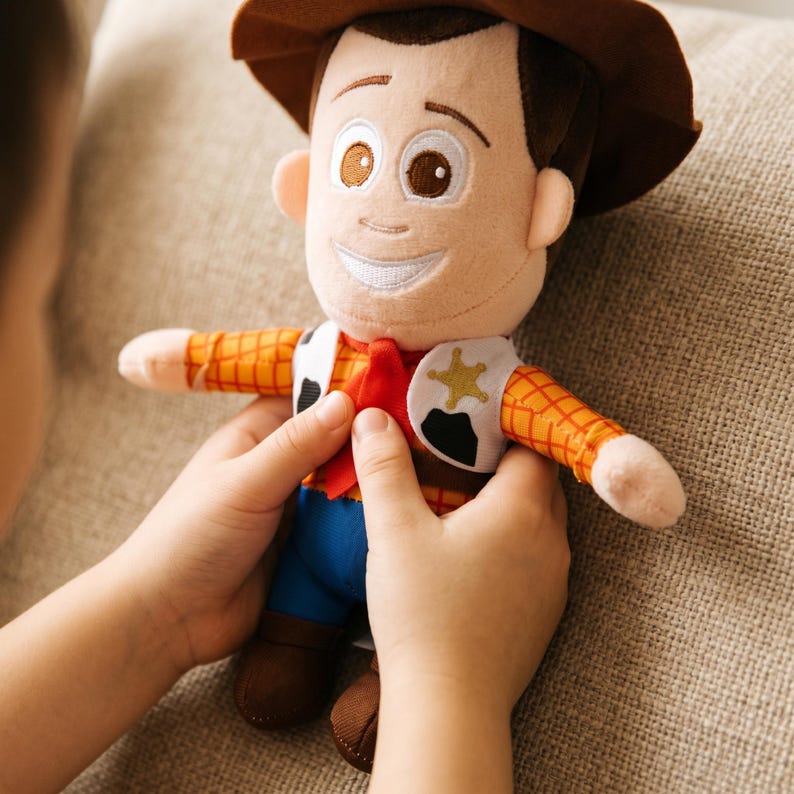 May include: A plush doll of a cowboy character featuring a brown hat, a yellow and orange plaid shirt, a red neckerchief, a white vest with a gold star emblem, blue pants, and brown boots. The doll has embroidered facial features and brown hair.