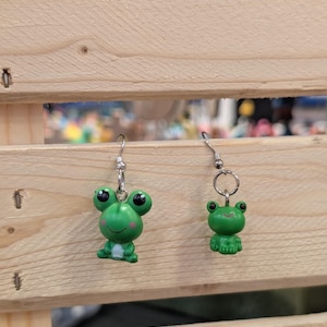 May include: A pair of mismatched dangle earrings featuring small green frog charms. The left earring displays a larger frog with black eyes and pink cheeks, while the right earring features a smaller, simpler green frog. Both hang from silver-toned metal hooks.