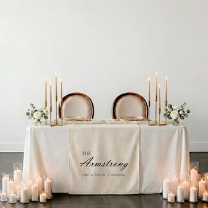 May include: A wedding table setting with a white tablecloth and a beige runner printed with "The Armstrong" and a date. Tall candles and floral arrangements are placed on the table, creating a romantic atmosphere.