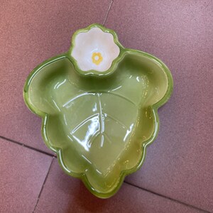 May include: A green ceramic leaf-shaped serving dish with a small flower-shaped bowl attached. The leaf dish has a glossy finish with a detailed vein pattern. The flower bowl is white with a yellow center. A decorative piece for serving snacks.