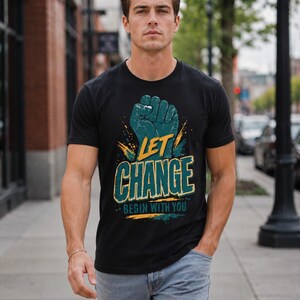 May include: A black short-sleeved t-shirt featuring a graphic of a teal raised fist above the yellow text "LET CHANGE" and the smaller teal text "BEGIN WITH YOU," accented with yellow and teal paint splatter effects on the front.