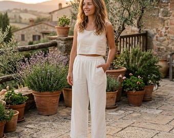 May include: A woman wearing a cream-colored linen crop top and wide-leg pants. The outfit is simple and elegant, perfect for warm weather. The pants have pockets. The background features terracotta pots with plants, a stone wall, and a scenic view.