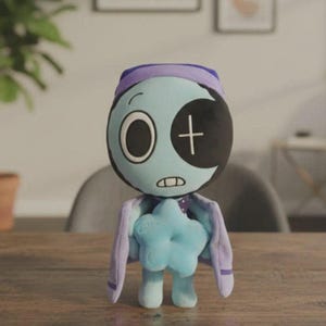 Puede incluir: Muñeco de peluche con una cara azul claro, un ojo negro en forma de media luna y una cruz blanca. Lleva un sombrero y una capa morados y sostiene una estrella azul. El muñeco está sobre una mesa de madera.