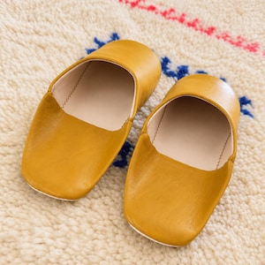 Puede incluir: Un par de zapatillas de cuero color amarillo mostaza con diseño de punta redondeada, acabado exterior liso y forro interior en color beige claro, con costuras visibles a lo largo de los bordes de la abertura y la suela.