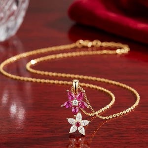 Puede incluir: Un collar de cadena dorada con un colgante de dos flores superpuestas. La flor superior tiene cinco piedras rosas de corte marquesa y una piedra central clara, mientras que la inferior tiene cinco piedras claras de corte marquesa y una piedra central rosa.