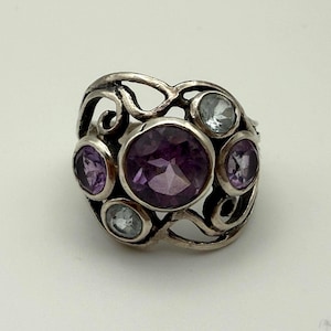 May include: A silver ring featuring a large central round purple amethyst gemstone surrounded by two smaller round purple amethysts and two light blue round gemstones, all set within an openwork metal band with swirling, organic metal patterns.
