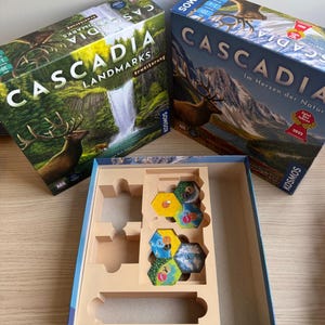 Könnte beinhalten: Zwei Brettspielschachteln mit den Titeln Cascadia Landmarks und Cascadia Im Herzen der Natur, die Elche und Landschaften zeigen. Vorne steht eine geöffnete Spielschachtel mit einem hellen Kunststoffeinsatz und sechseckigen Plättchen.