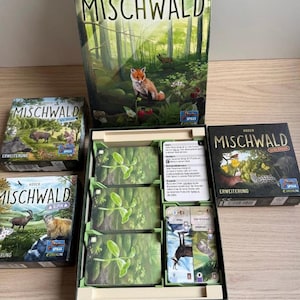 Könnte beinhalten: Ein Mischwald Brettspiel-Set bestehend aus einer großen vertikalen Spielschachtel, zwei kleineren Erweiterungsboxen und einer offenen Spielschachtel mit drei Kartenstapeln mit grünen Blattmotiven, einer Spielanleitung und einer Reihe Spielkarten.