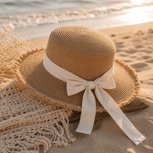 May include: A brown straw sun hat with a wide brim and a cream-coloured ribbon tied in a bow. The hat is resting on a sandy surface near the ocean. The hat's brim has a frayed edge.