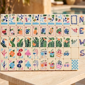 May include: A set of cream-colored mahjong tiles arranged in a grid. The tiles feature colorful illustrations of tropical fruits, flowers, beach items, and numbers one through nine. Some tiles display the word JOKER and various summer-themed icons.