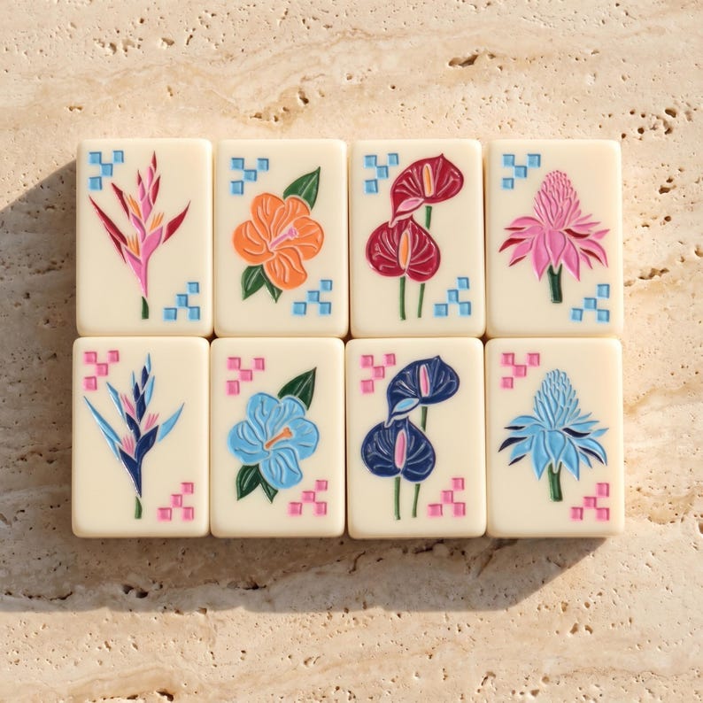 May include: Eight rectangular off-white mahjong tiles arranged in two rows. Each tile features a colorful engraved tropical flower design, including hibiscus, anthurium, and ginger, accented by small geometric square patterns in blue or pink.