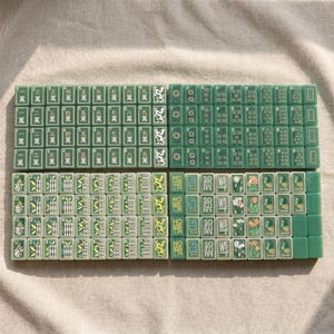 May include: A set of green Mahjong tiles arranged in four rectangular blocks. The tiles feature various designs including Chinese characters, numeric dots, bamboo patterns, directional text like North, South, East, West, and floral illustrations.