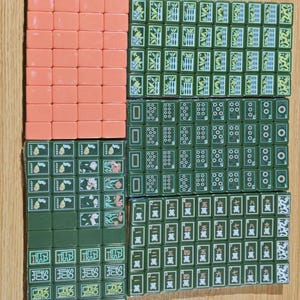 May include: A collection of rectangular mahjong tiles arranged in a grid. The set includes solid peach-colored tiles and dark green tiles featuring various bamboo patterns, circular designs, Chinese characters, joker labels, and directional markers.
