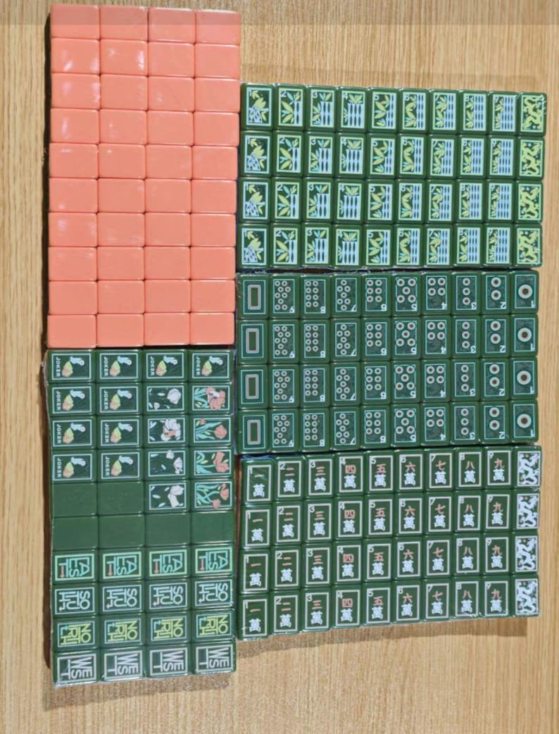 May include: A collection of rectangular mahjong tiles arranged in a grid. The set includes solid peach-colored tiles and dark green tiles featuring various bamboo patterns, circular designs, Chinese characters, joker labels, and directional markers.