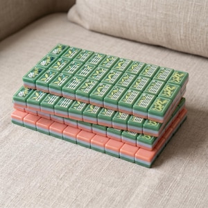 May include: A set of rectangular mahjong tiles stacked in four rows. The top tiles feature green surfaces with white bamboo, numerical, and dragon engravings. Each tile has a layered design with a green middle section and a pink base.
