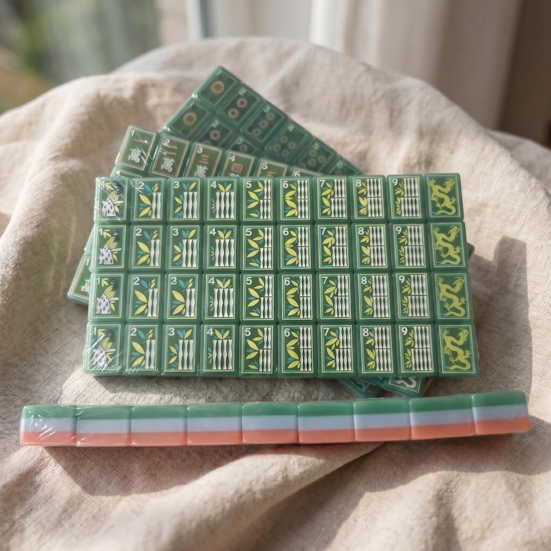 May include: A set of green Mahjong tiles featuring bamboo, dragon, and numerical designs in white, yellow, and black. The tiles are arranged in a rectangular grid with a single row of tiles placed horizontally in front, showing a tri-color side profile.