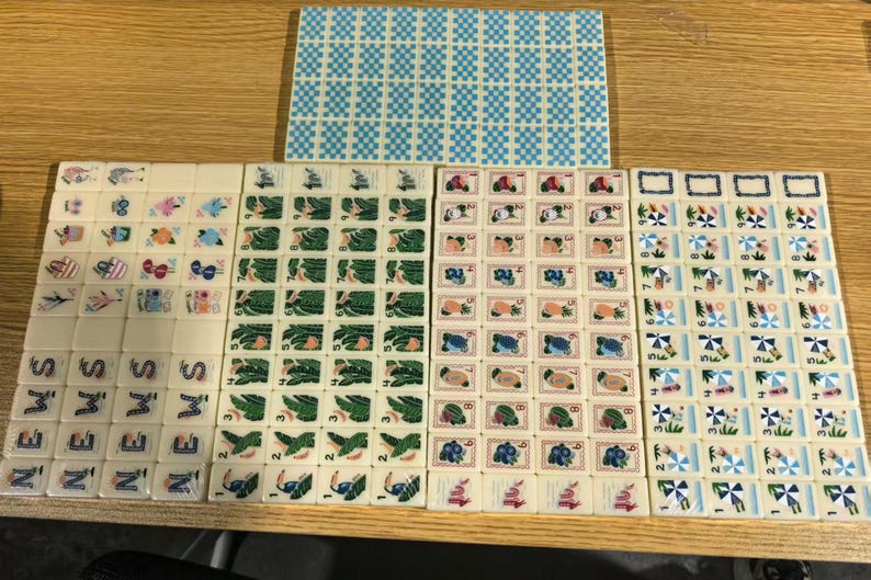 May include: A collection of rectangular mahjong tiles arranged in rows and columns. The tiles feature various colorful illustrations including tropical plants, fruits, beach umbrellas, and the word NEWS. One large rectangular tile has a blue checkered pattern.