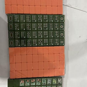 May include: A set of rectangular Mahjong tiles arranged in four rows. Two rows consist of solid peach-colored tiles, while the other two rows feature dark green tiles with white numerical markings, floral illustrations, and text including JOKER, SOUTH, NORTH, and WEST.