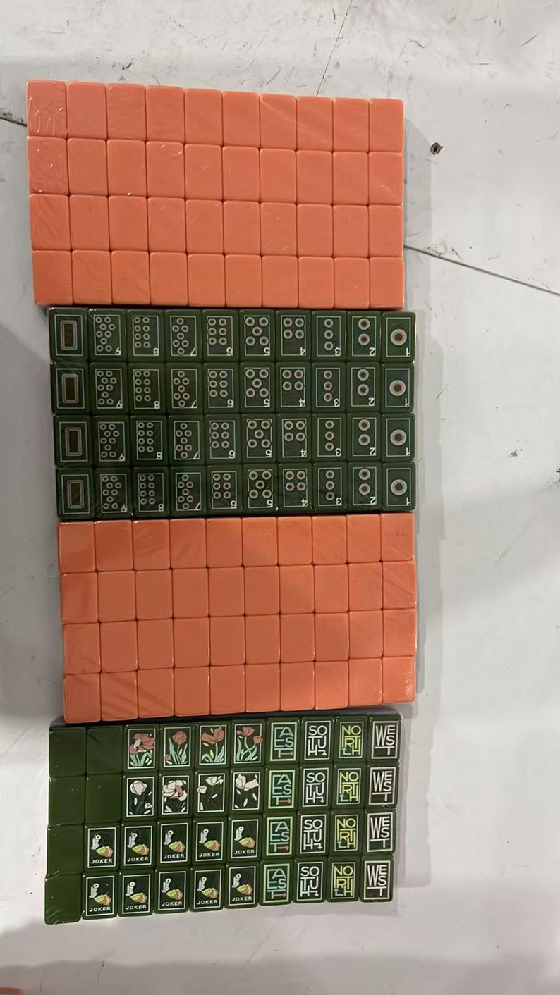 May include: A set of rectangular Mahjong tiles arranged in four rows. Two rows consist of solid peach-colored tiles, while the other two rows feature dark green tiles with white numerical markings, floral illustrations, and text including JOKER, SOUTH, NORTH, and WEST.