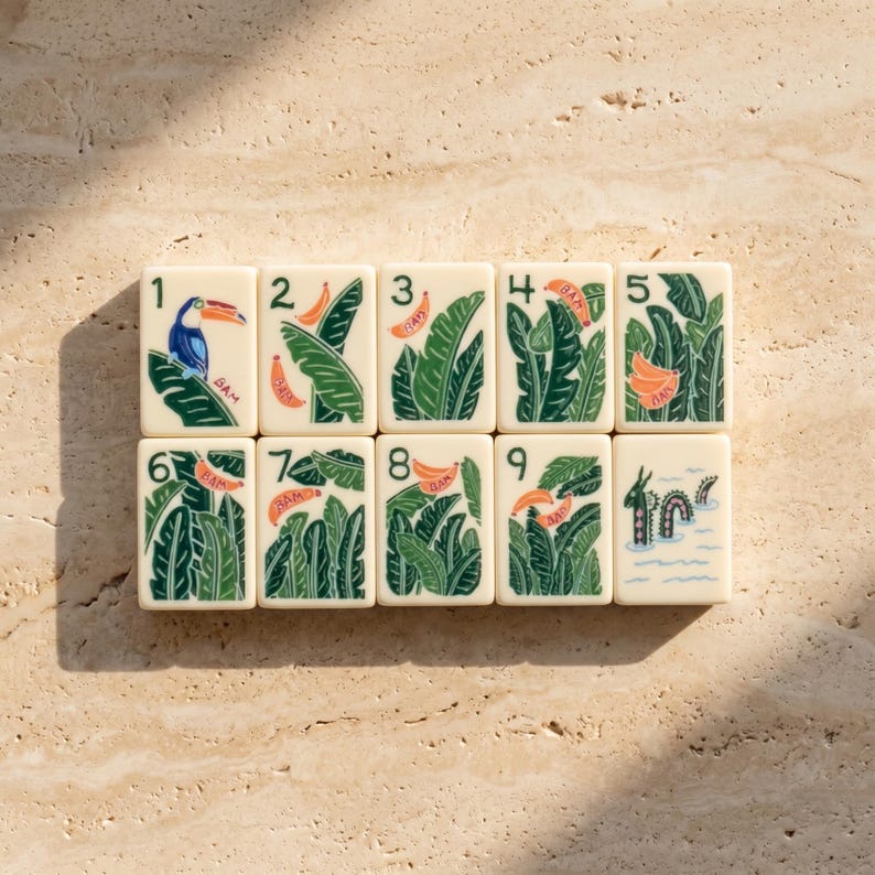 May include: A set of ten rectangular off-white tiles arranged in two rows, numbered one through nine plus a tenth tile. Each tile features green tropical leaf illustrations, orange banana motifs, a blue toucan on tile one, and a sea creature on tile ten.