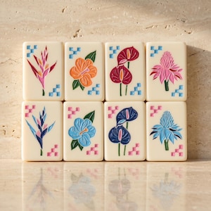 May include: Eight rectangular off-white tiles arranged in two rows, each featuring a colorful engraved tropical flower illustration and small blue or pink geometric corner accents. The designs include bird of paradise, hibiscus, and anthurium flowers.