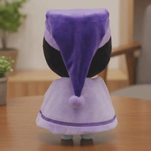 Peut inclure: Poup&eacute;e en peluche v&ecirc;tue d'un bonnet de nuit et d'une cape violets. Le bonnet de nuit est violet fonc&eacute;, et la cape est d'une nuance de violet plus claire avec une bordure blanche. La poup&eacute;e a une t&ecirc;te noire et de petits pieds bleus.