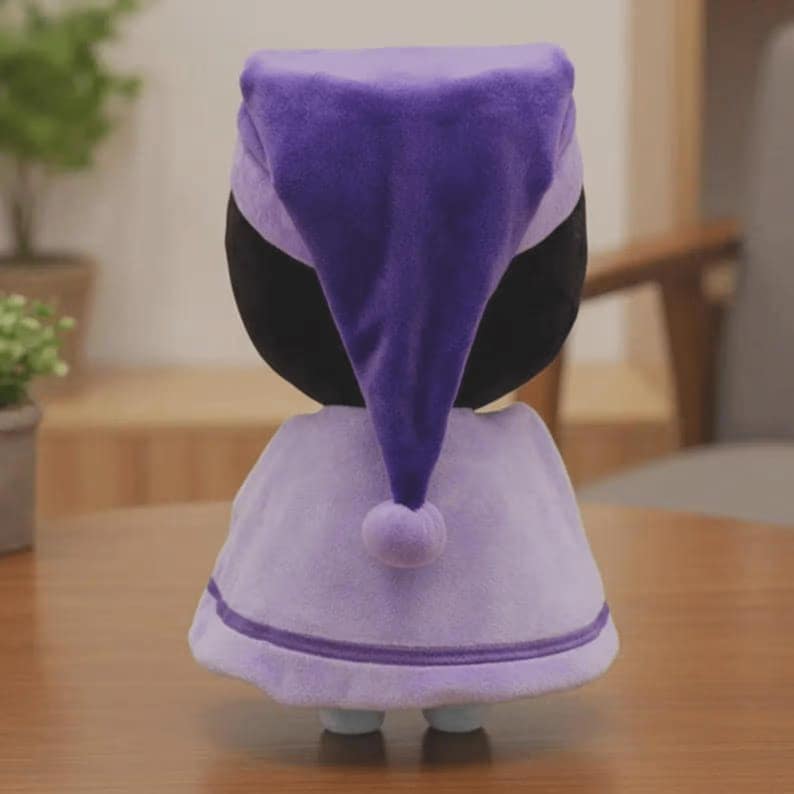 Peut inclure: Poup&eacute;e en peluche v&ecirc;tue d'un bonnet de nuit et d'une cape violets. Le bonnet de nuit est violet fonc&eacute;, et la cape est d'une nuance de violet plus claire avec une bordure blanche. La poup&eacute;e a une t&ecirc;te noire et de petits pieds bleus.