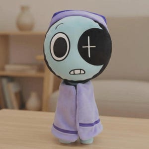 Peut inclure: Une poup&eacute;e en peluche avec un visage bleu clair, un grand &oelig;il et un patch noir avec une croix blanche. Elle porte une casquette et une cape violettes. La poup&eacute;e a une petite bouche cousue et est debout.