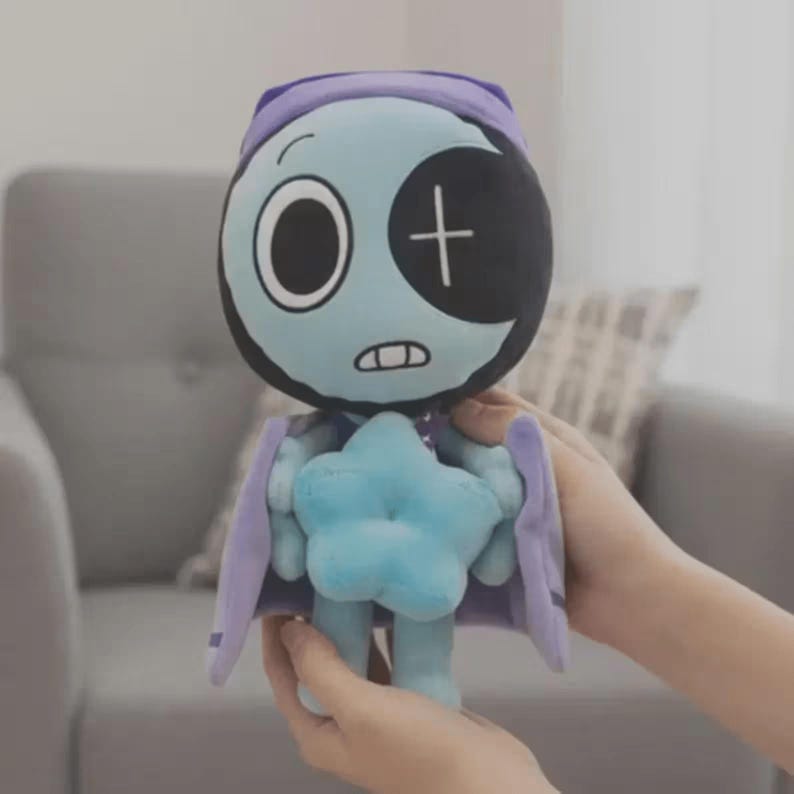 Peut inclure: Une poup&eacute;e en peluche avec un visage bleu clair, un cache-&oelig;il noir en forme de croissant et un bonnet de nuit violet. La poup&eacute;e a un corps en forme d'&eacute;toile bleu clair et des ailes en forme de cape violette. La poup&eacute;e est tenue par une personne.