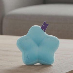 Peut inclure: Un jouet en peluche en forme d'&eacute;toile bleu clair avec des d&eacute;tails cousus. Un ruban violet avec des croix blanches est ins&eacute;r&eacute; dans le haut de l'&eacute;toile. Le jouet est pos&eacute; sur une surface en bois.
