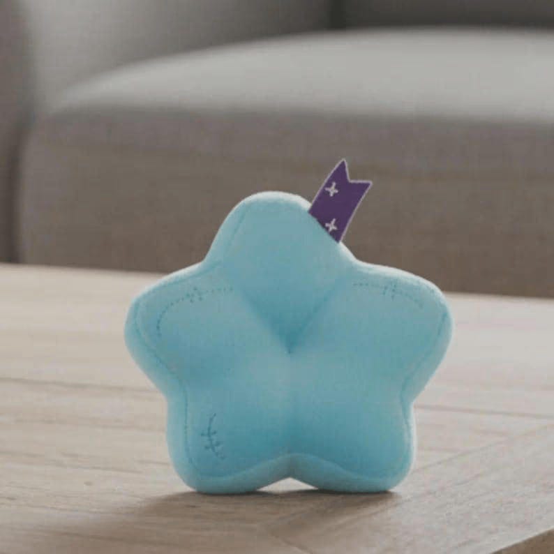Peut inclure: Un jouet en peluche en forme d'&eacute;toile bleu clair avec des d&eacute;tails cousus. Un ruban violet avec des croix blanches est ins&eacute;r&eacute; dans le haut de l'&eacute;toile. Le jouet est pos&eacute; sur une surface en bois.
