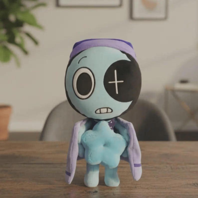 Peut inclure: Une poup&eacute;e en peluche avec un visage bleu clair, un cache-&oelig;il noir en forme de croissant et une croix blanche. Elle porte un chapeau et une cape violets et tient une &eacute;toile bleue. La poup&eacute;e a une bouche cousue et un seul &oelig;il.