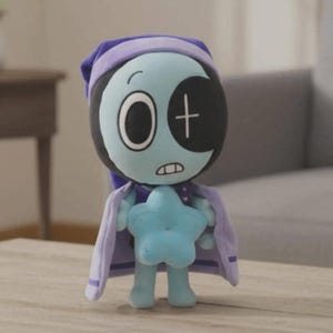 Peut inclure: Poup&eacute;e en peluche avec un visage bleu clair, de grands yeux et un cache-&oelig;il noir avec une croix blanche. Elle porte un bonnet de nuit et une cape violets et tient une &eacute;toile bleue. La poup&eacute;e est sur une surface en bois.