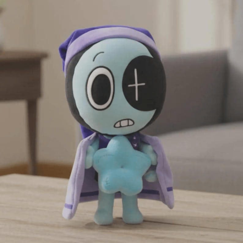 Peut inclure: Poup&eacute;e en peluche avec un visage bleu clair, de grands yeux et un cache-&oelig;il noir avec une croix blanche. Elle porte un bonnet de nuit et une cape violets et tient une &eacute;toile bleue. La poup&eacute;e est sur une surface en bois.