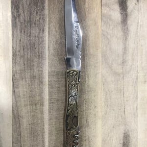 May include: A stainless steel knife with a pointed blade and a decorative handle. The handle features an engraved fish design and the text "Fightin' Fish". The knife is set against a wooden background.