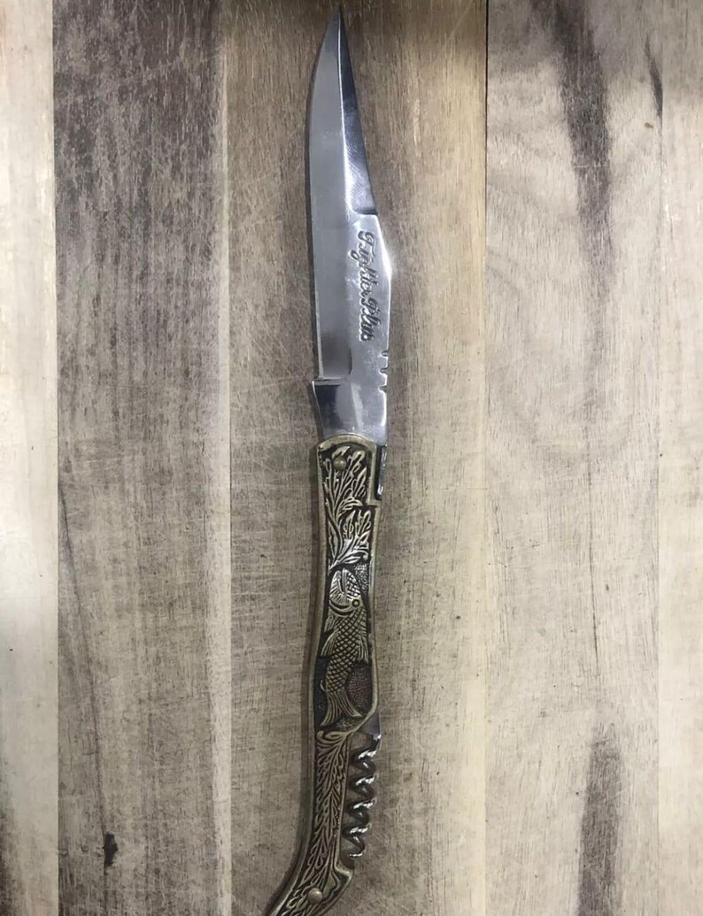May include: A stainless steel knife with a pointed blade and a decorative handle. The handle features an engraved fish design and the text "Fightin' Fish". The knife is set against a wooden background.