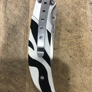 K&ouml;nnte beinhalten: Ein Klappmesser mit Zebrastreifenmuster am Griff. Der Griff ist wei&szlig; mit schwarzen Streifen. Es hat einen silbernen Metall-Taschenclip und den Text "MODEL 776".