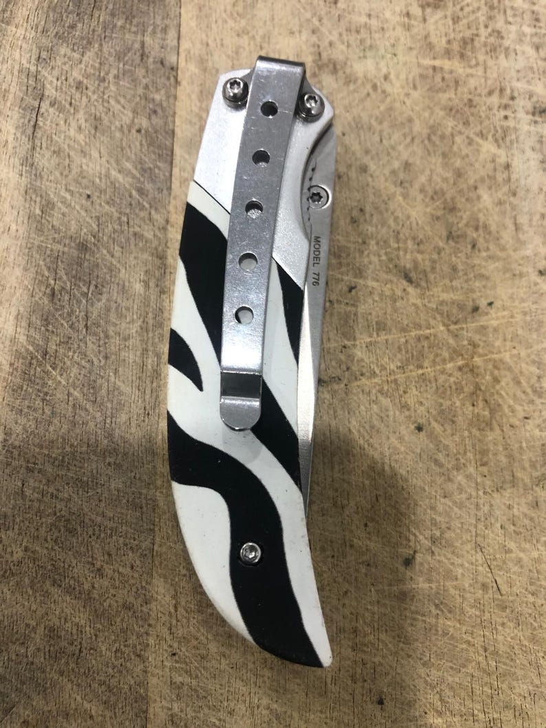 K&ouml;nnte beinhalten: Ein Klappmesser mit Zebrastreifenmuster am Griff. Der Griff ist wei&szlig; mit schwarzen Streifen. Es hat einen silbernen Metall-Taschenclip und den Text "MODEL 776".