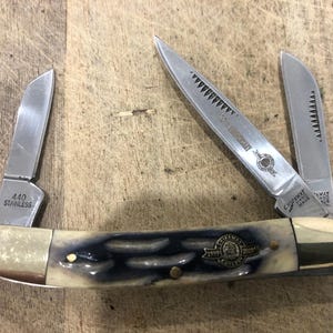 Op de afbeelding: Een zakmes met meerdere messen met een crème en blauw handvat. De roestvrijstalen messen zijn gedeeltelijk open en tonen de tekst "440 Stainless" en "125th Anniversary". Het mes ligt op een houten oppervlak.