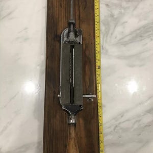 May include: An antique knife sharpener mounted on a wooden base. The sharpener is made of metal with a wooden handle. A yellow measuring tape is placed next to the sharpener, showing a length of approximately 24 inches.
